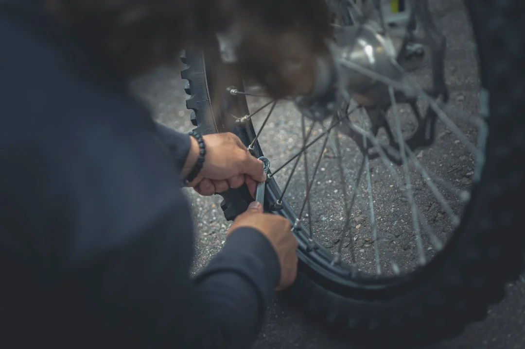 A gravel cyclist kneeling trailside, inserting a tubeless plug into a rear tire with a CO₂ inflator and patch kit laid out on the ground nearby